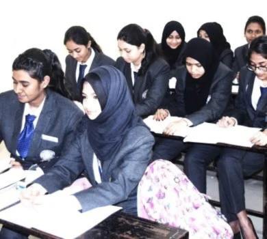 Female students in uniform sitting at desks in a classroom, listening attentively and taking notes.