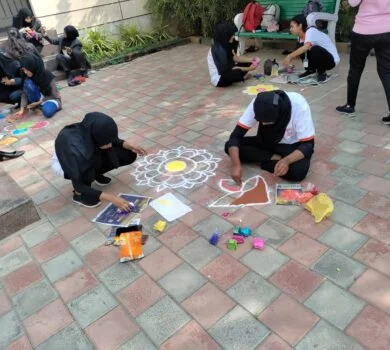 Two students drawing and coloring a detailed rangoli pattern on the pavement.