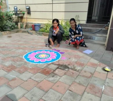Two students creating a large, vibrant floral rangoli design outdoors in the courtyard.