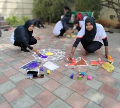 Students sitting on the ground outside, working collaboratively on a colorful rangoli.