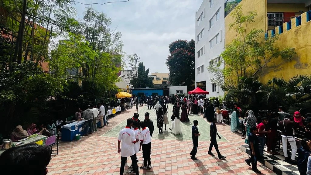 An outdoor courtyard scene with a crowd of students, food stalls under a red tent, and white balloons.